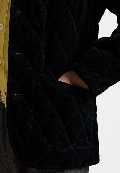 Melnas korduroja pledu jaka ar dimanta rakstu un priekšējo kabatu, kompleksā apģērba kombinācijā ar dzelteno pogājamo krekli. Ievērojami ir teksturētā auduma un dizaina detaļas.