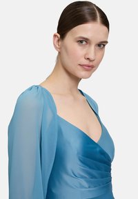 Une robe ajustée en satin lisse de couleur bleu clair, avec des manches bouffantes transparentes et un décolleté plongeant ; froncée à la taille.