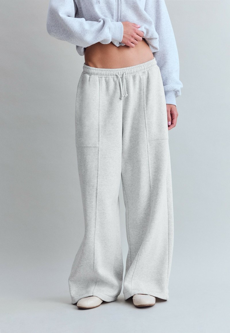Person wearing light gray wide-leg sweatpants, cropped light gray sweatshirt, and white shoes, standing against a plain gray background.