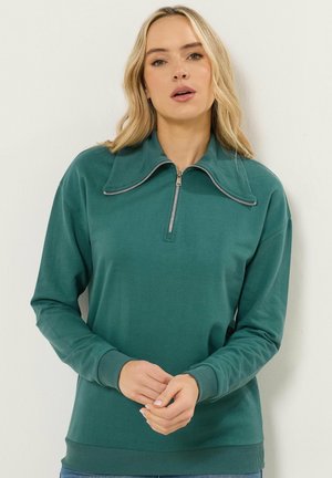 Blonde vrouw die een tealkleurige trui met lange mouwen, een brede kraag en een halve ritssluiting draagt, staand tegen een lichte, effen muur.