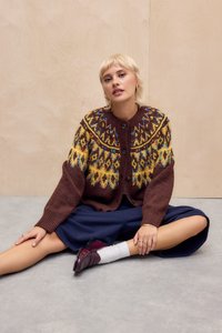 Brūns adīts kardigans ar krāsainām ģeometriskām rakstām, oversized piegriezums, pogu aizdare, uzvilkts pār tumši zilu svārku. Modelis sēž uz grīdas.
