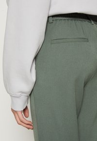 Groene broek met een textuurstof, voorzien van een elastische tailleband en een enkele achterzak. Een lichtgrijze mouw is zichtbaar.