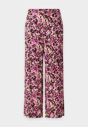 Femme portant un haut blanc côtelé à manches courtes, un pantalon large à fleurs rose et noir, et des talons noirs pointus, debout avec une main dans la poche.