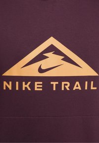 Sweat-shirt bordeaux avec un grand graphique triangulaire doré comportant un logo Nike stylisé et les mots "NIKE TRAIL" en lettres majuscules.
