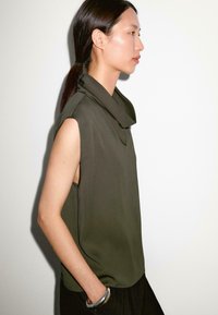 Top senza maniche di colore verde oliva con colletto drappeggiato, tessuto morbido e spacchi laterali. Caratterizzato da una vestibilità comoda e una superficie liscia.