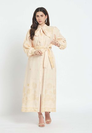 Ženska, oblečena v dolgo, bledo breskovo obleko z rožnatimi vzorci, dolgimi napihnjenimi rokavi, zavezanim pasom in pentljo ob ovratniku, stoji pred belim ozadjem.