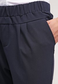 Pantalons bleu marine avec une ceinture élastique, dotés de plis avant et de poches latérales. Fabriqués en tissu lisse et léger.
