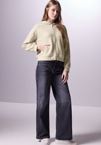 Sudadera de cremallera color verde claro con textura, capucha y bolsillo frontal, combinada con pantalones vaqueros de pierna ancha de mezclilla oscura y zapatillas blancas.