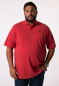 Camisa polo roja de manga corta, con cuello y un pequeño logotipo azul marino en el lado izquierdo del pecho. Lleva un par de vaqueros azul oscuro.