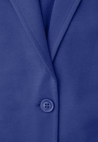 Gros plan d'un blazer bleu avec un seul bouton fermé, montrant les détails des coutures et la texture du tissu.