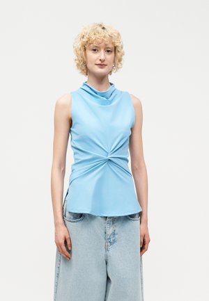 Νεαρό άτομο με κοντά σγουρά ξανθά μαλλιά φοράει αμάνικη ανοιχτό μπλε μπλούζα με στριφτό ντεκολτέ και φαρδιά ανοιχτό τζιν παντελόνια σε λευκό φόντο.