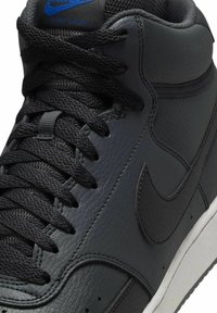 Zapatilla de baloncesto alta de cuero negro con acabado texturizado, que presenta una suela blanca y cordones negros a juego. Logo azul de Nike en la lengüeta.