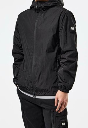 Mand iført sort hættetrøje med lynlås og sorte cargobukser, hænder afslappet ned langs siderne mod hvid baggrund.