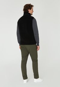 Svart fleeceväst med hög krage, ihopparad med en grå långärmad tröja och olivgröna byxor; modellen vänder sig bort, iklädd vita sneakers.