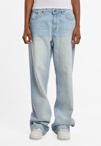 Personne portant un jean bleu clair délavé et ample ainsi que des baskets blanches, debout avec les mains détendues le long du corps.