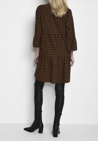 Braun-schwarz kariertes, gestuftes Kleid mit Dreiviertelärmeln, kombiniert mit schwarzen, figurbetonten Hosen und Stiefeletten. Glatte Stofftextur.