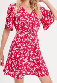Robe florale rouge avec un col en V, des manches courtes et évasées, et une taille empire. Présente des motifs de fleurs blanches et une jupe évasée. Tissu doux.