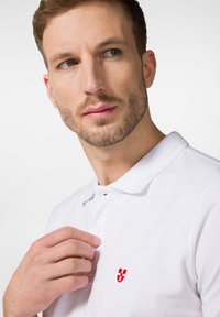 Biała koszulka polo z krótkim kołnierzykiem, dwoma guzikami i małym, czerwonym, haftowanym logo po lewej stronie. Gładka bawełniana struktura.