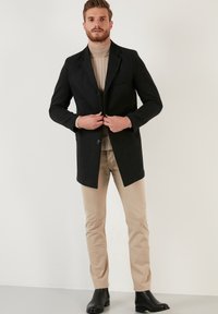 Abrigo de mezcla de lana negra con un diseño entallado, cierre de un solo botón y solapas de muesca, combinado con un suéter de cuello alto beige y pantalones beige ajustados.