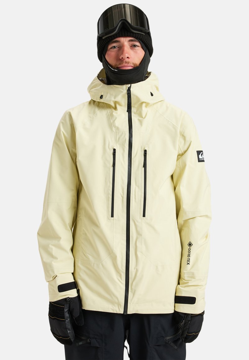 Gelbe wasserdichte Jacke mit hohem Kragen, schwarzen Reißverschlusstaschen und GORE-TEX-Branding. Getragen mit schwarzen Handschuhen und einer Skimütze.