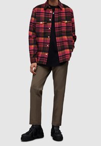 Czerwona, różowa i czarna koszula flanelowa w kratę z dwiema kieszeniami z przodu, założona na czarną koszulę. W zestawie z beżowymi spodniami i czarnymi butami na sznurowadła.