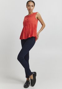 Rød, ærmeløs bluse med struktureret design og knapdetalje ved halsen, parret med mørkeblå skinny jeans og sorte loafers.