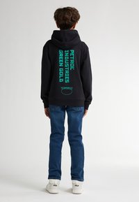 Sudadera negra con un texto en negrita color verde azulado que dice "PETROL INDUSTRIES GREEN GOLD" en la espalda, combinada con unos vaqueros de denim azul y zapatillas blancas.