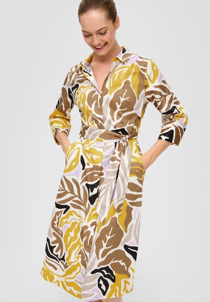 Robe chemise avec un col, présentant un imprime de feuillage audacieux en jaune, marron et noir sur un fond clair. Comprend une taille cintrée avec un nœud et des poches.