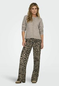 Lichtgrijze gebreide cardigan met knopen, gecombineerd met beige broeken met een luipaardprint en wijde pijpen. Zachte textuur en ontspannen pasvorm. Bruine schoenen zichtbaar.