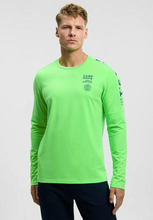 Mann trägt hellgrünes langärmeliges Shirt mit blauem Text auf Brust und Ärmel, kombiniert mit dunkler Hose, vor einfarbigem Hintergrund.