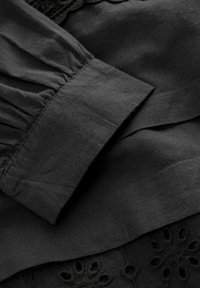 Tela negra con superficie texturizada, con mangas fruncidas y acentos de bordado floral. Textura suave con elementos de diseño en capas.