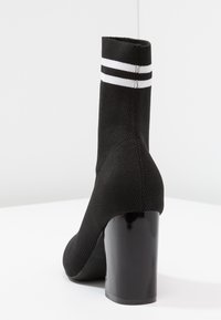Bottine noire avec un dessus en tricot côtelé, des rayures blanches en haut et un large talon noir brillant. Texture lisse et forme épurée.