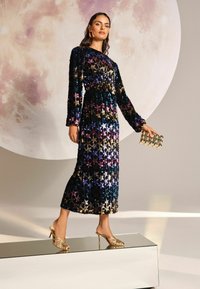 Földig érő, hosszú ujjú fekete ruhát multicolor csillag paillettekkel díszítve. Arany sarkú szandálokkal és egy hozzá illő arany clutch táskával párosítva.