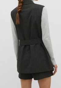 Gilet grigio scuro a righe con cintura, indossato sopra una camicia a maniche lunghe grigio chiaro, abbinato a pantaloni corti coordinati. Tessuto texturizzato.