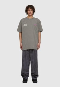 Pelēks pārspīlēts T-krekls ar baltu un rozā tekstu, apvienots ar brīvām pelēkām biksēm. Audums izskatās mīksts un ar brīvu piegriezumu. Melni kedas.