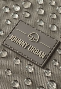 Textúrázott szürke anyag vízcseppekkel; egy szürke bőrcímke látható, amelyen "JOHNNY URBAN" van begyomva. Mintás felület tiszta, fényes kiemelésekkel.