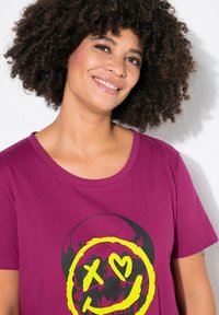 Lilla bomulds T-shirt med et stort sort kraniegrafik, der har et gult smilende ansigt, hjerteøjne og tykke konturer. Korte ærmer og rund halsudskæring.