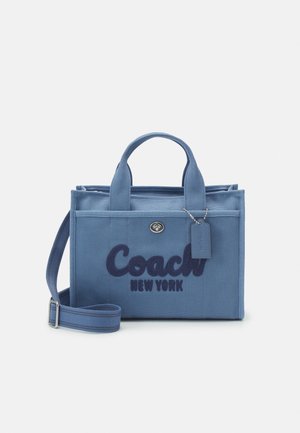 Sac à main en tissu bleu avec deux poignées et une bandoulière réglable, avec le logo "Coach New York" et une petite étiquette attachée.