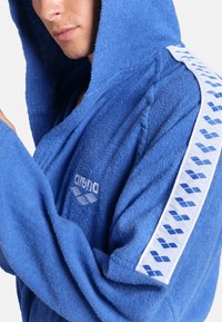 Blauwe badjas met capuchon, gemaakt van badstof. Kenmerkt zich door een witte zijstrip met blauwe logo's en een gestructureerde stof. Inclusief een logo-detail aan de voorkant.