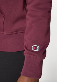 Sweatshirt bordeaux avec poignets côtelés, arborant le logo de Champion brodé en blanc et rouge sur la manche, fabriqué à partir d'un tissu en mélange de coton doux.