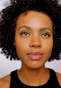 Portret van een persoon met krullend, donker haar, die groene oogmake-up draagt en een septumpiercing heeft, tegen een neutrale achtergrond.
