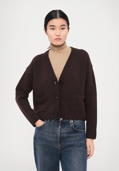 Brown wool cardigan with a v-neck, ribbed hems and cuffs, featuring two front pockets and black buttons. Worn over a beige turtleneck.