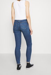 Jeans ajustados de color azul hechos de denim, con una cintura alta, cinco bolsillos y costuras sutiles. Combinados con zapatillas negras de caña alta.