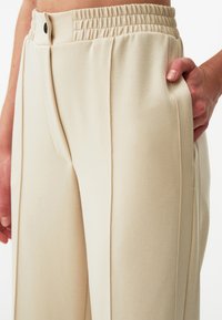 Beżowe spodnie o szerokich nogawkach, wykonane z miękkiego materiału, z fakturowanym elastycznym pasem i funkcjonalnymi bocznymi kieszeniami z lamówką.