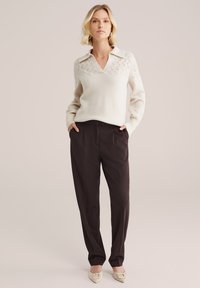 Krem pulover s pletenjem in V-izrezom ter detajlom ovratnika, v kombinaciji z visokim pasom, temno rjavimi hlačami in svetlimi čevlji s koničastim nosom.