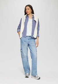 Tenue décontractée composée d'une chemise bleue à manches longues, d'un gilet en laine crème, d'un t-shirt rayé blanc, d'un pantalon cargo bleu clair et de baskets argentées.
