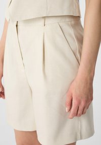 Beige tailored shorts with pleats, made from textured fabric. Features a mid-rise waist and side pockets for functional styling.