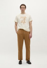 Romantisch t-shirt in crème met een oranje grafische print en korte mouwen, gecombineerd met lichtbruine, luchtige recht toegesneden broeken en donkere schoenen.