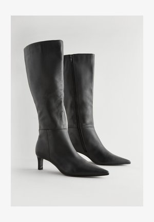 Botas negras de cuero hasta la rodilla con punta afilada, tacón bajo en bloque y cremallera lateral. La superficie tiene una textura lisa con pocos detalles.