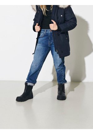 Bottines noires imperméables, jeans bleu légèrement usés et retroussés, et une veste sombre avec une capuche en fourrure. Design simple et fonctionnel.